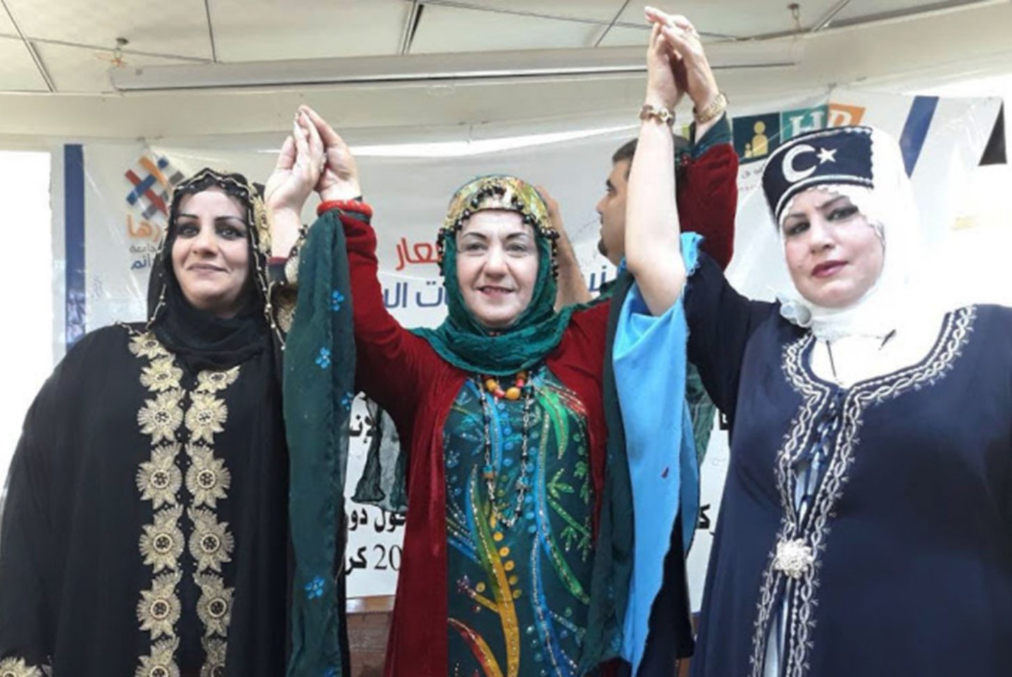 Photo of three Iraqi women from different ethnic backgrounds in traditional dress raising their hands together in solidarity.